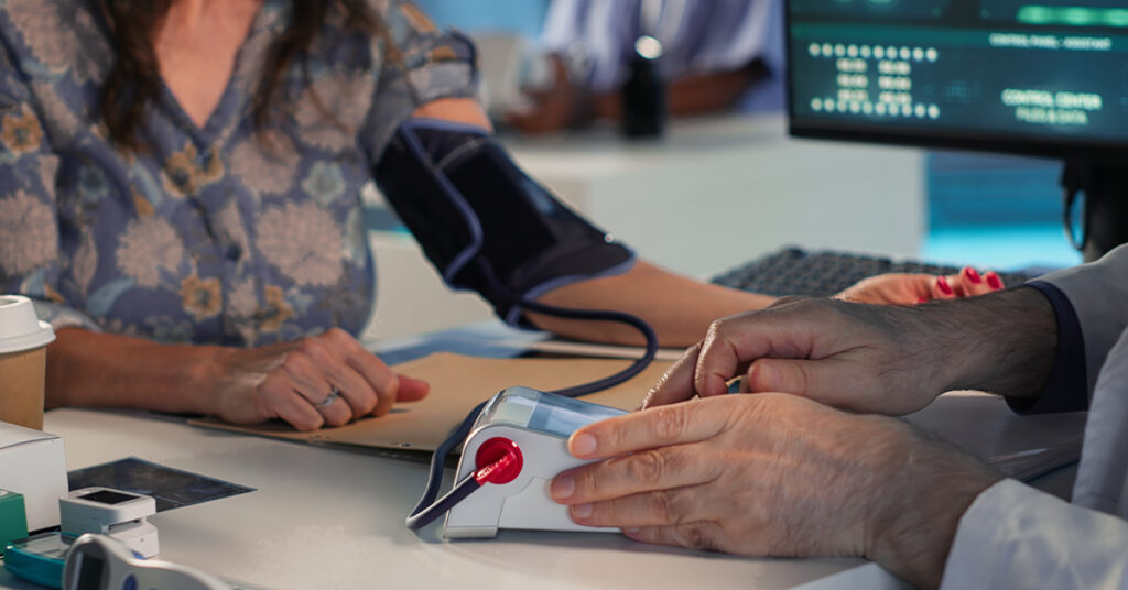 A close-up of a doctor's hands operating a digital monitor while checking a patient's blood pressure with an arm cuff.
