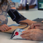 A close-up of a doctor's hands operating a digital monitor while checking a patient's blood pressure with an arm cuff.