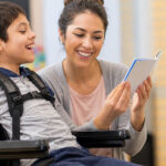 Boy in wheelchair reading with a smiling caregiver.