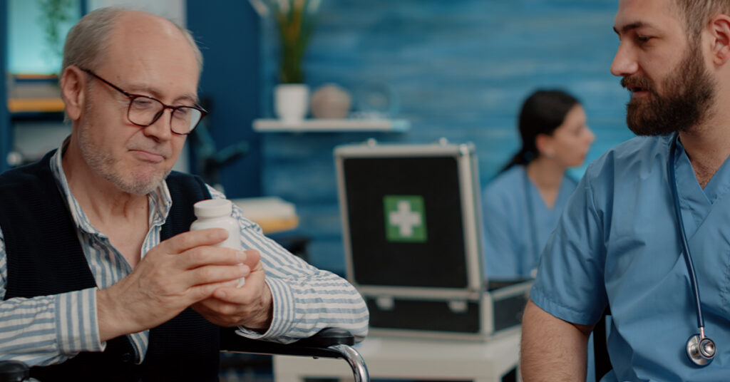 An elderly man in a wheelchair reviews a pill bottle with a healthcare worker in a clinical setting.