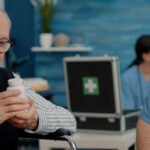 An elderly man in a wheelchair reviews a pill bottle with a healthcare worker in a clinical setting.