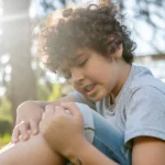 Child sitting outdoors and holding a painful knee.